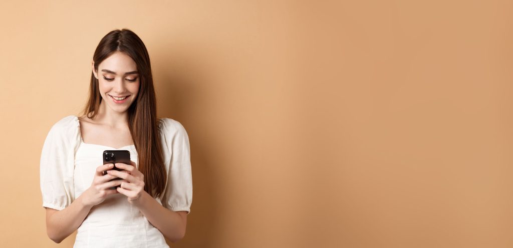 messaggio di testo della giovane donna sul telefono cellulare che sorride e che legge lo schermo dello smartphone che sta sul beige